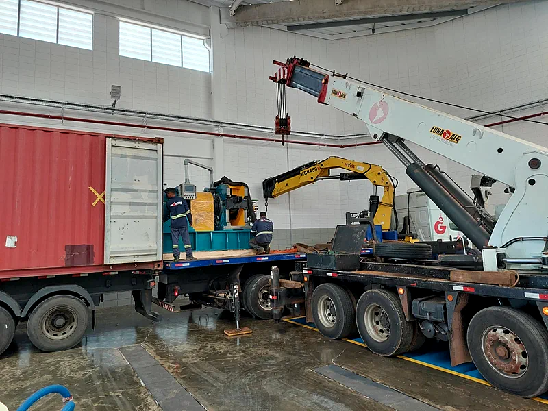 Dois operadores de uniforme acompanhando o carregamento de uma máquina em carreta prancha, dentro de um galpão
