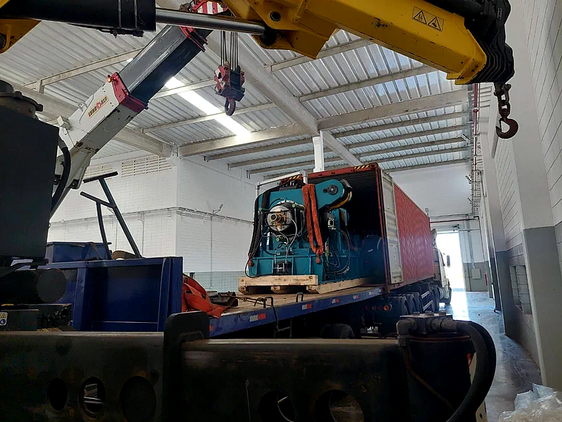 Guindaste articulado içando um equipamento industrial para dentro de um contêiner apoiado sobre carreta prancha