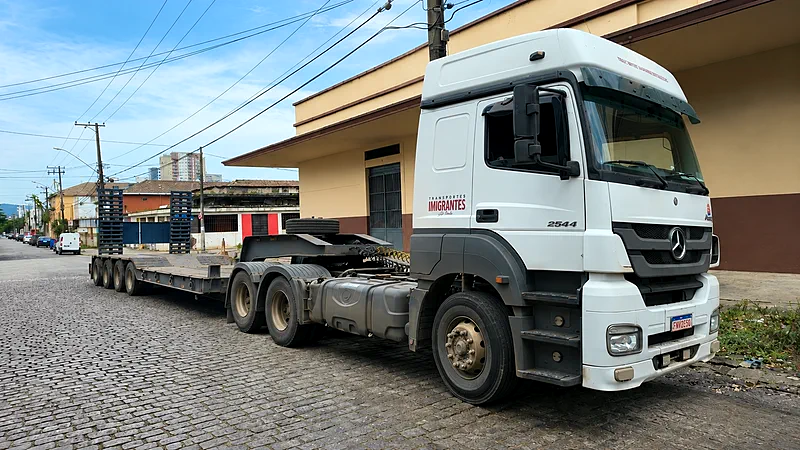 Caminhão Mercedes-Benz Actros branco da Transportes Imigrantes engatado a uma carreta prancha de três eixos, estacionado em rua de paralelepípedos