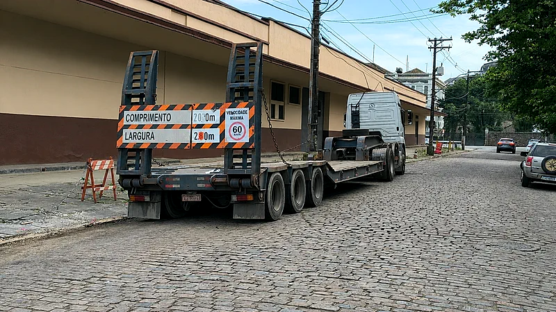 Traseira de carreta prancha com placa de sinalização de comprimento e largura para carga excedente, trafegando em rua de paralelepípedos