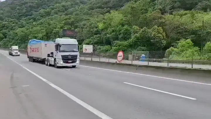 Caminhão branco com contêiner de 40 pés sobre a carroceria, trafegando por rodovia cercada de mata, com outro caminhão ao fundo