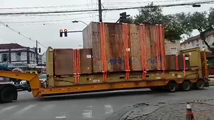 Carreta extensível com caixas de madeira amarradas manobrando em via urbana, acompanhada por um operador em solo