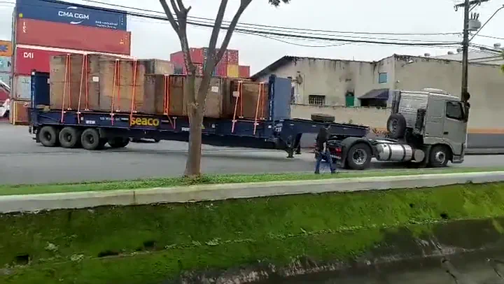 Carreta prancha estendida carregada com estruturas de madeira amarradas, passando em frente a um pátio de contêineres empilhados